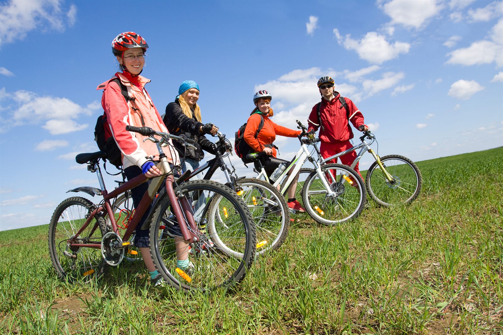 Four cyclists in helmets standing with their mountain bikes on a grassy hillside under a blue sky